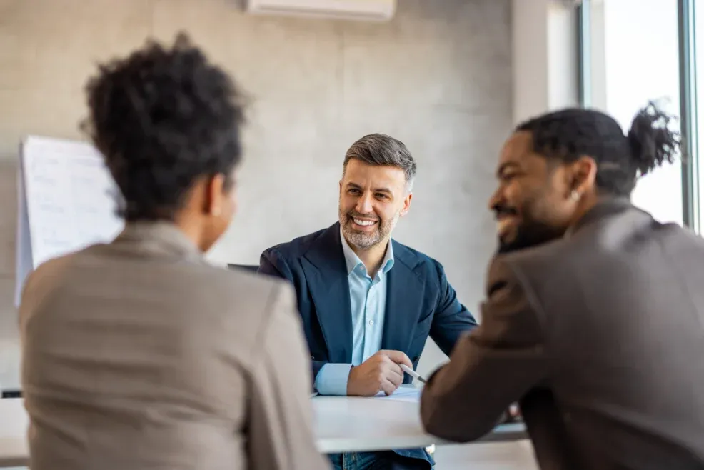 A smiling attorney in a navy blazer meets with two clients across a table in a bright office.