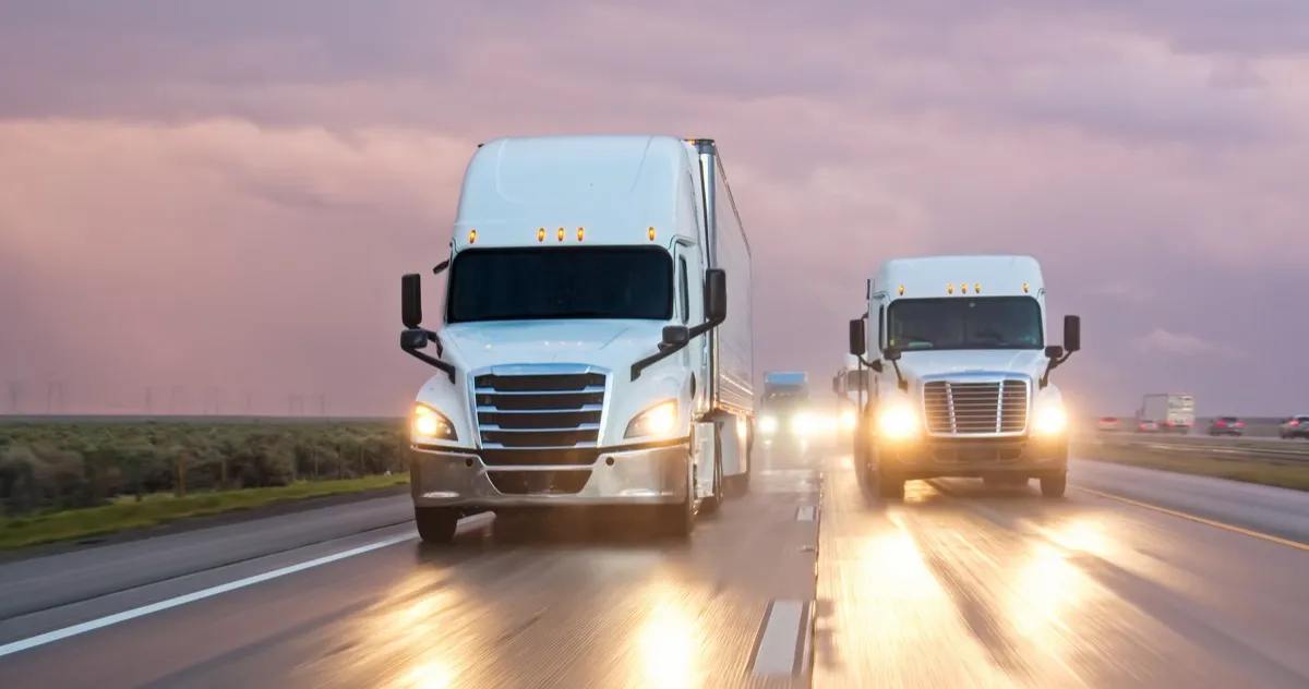 Two semi trucks with headlights on driving down a highway at dusk.