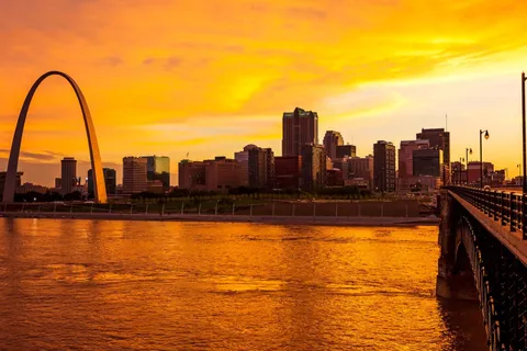 The Gateway Arch and downtown St. Louis skyline at sunset, seen across the Mississippi River from the Eads Bridge.